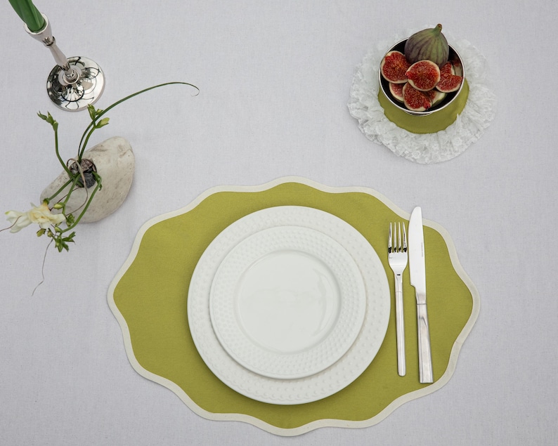 Op de afbeelding: Bovenaanzicht van een tafelsetting met een groene placemat, witte borden en bestek. Een kleine kom met vijgen staat op een kanten kleedje. Een kleine vaas met bloemen is ook aanwezig.