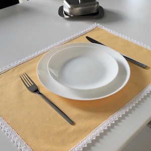 Può includere: Un'apparecchiatura da tavola con un runner giallo bordato di pizzo bianco, un piatto e una ciotola bianchi, posate e una burriera in acciaio inossidabile. Forchetta, coltello e burriera sono in acciaio inossidabile.