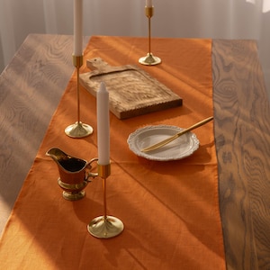 Könnte beinhalten: Eine Nahaufnahme einer Tischdekoration mit einem braunen Holztisch, einem orangefarbenen Tischläufer aus Leinen und drei goldenen Kerzenständern mit weißen Kerzen. Auf dem Tischläufer befinden sich ein Holzbrett und ein weißer Teller mit einem goldenen Messer.