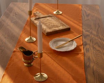 May include: A close-up of a table setting with a brown wooden table, an orange linen table runner, and three gold candle holders with white candles. There is a wooden cutting board and a white plate with a gold knife on the table runner.