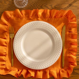 Puede incluir: Un plato blanco para cenar con una superficie texturizada se coloca sobre un mantel individual naranja con borde rizado. Un tenedor y un cuchillo dorados se colocan a la izquierda y a la derecha del plato.