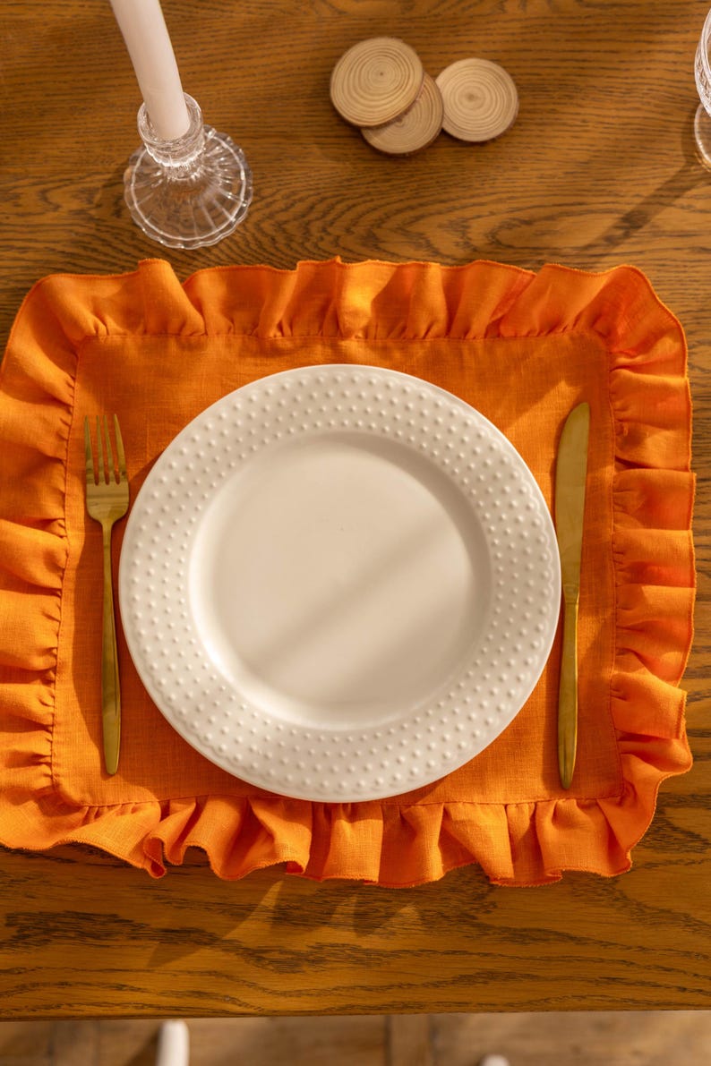 Puede incluir: Un plato blanco para cenar con una superficie texturizada se coloca sobre un mantel individual naranja con borde rizado. Un tenedor y un cuchillo dorados se colocan a la izquierda y a la derecha del plato.