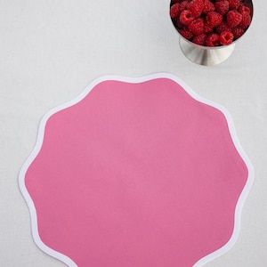May include: A bright pink, scalloped-edge placemat with a white border. A silver bowl filled with fresh red raspberries sits nearby. The placemat is on a white surface, creating a clean and inviting aesthetic.