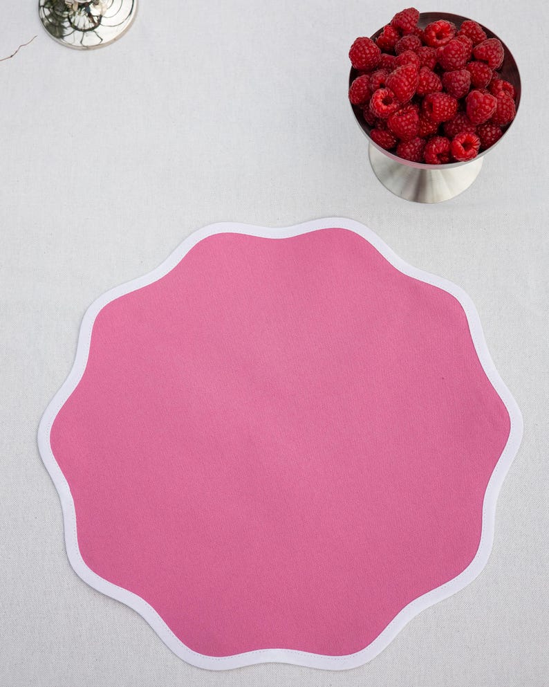 May include: A bright pink, scalloped-edge placemat with a white border. A silver bowl filled with fresh red raspberries sits nearby. The placemat is on a white surface, creating a clean and inviting aesthetic.