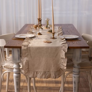 May include: A beige linen table runner with a ruffled edge, placed on a wooden table with two white chairs. The runner is decorated with two tall candlesticks and a small vase with white flowers.