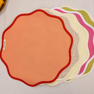 May include: A stack of colorful, scalloped placemats. The top placemat is peach with a red border. Other placemats include floral, yellow, pink, and green designs. The label on the peach placemat reads "COZY HOME".