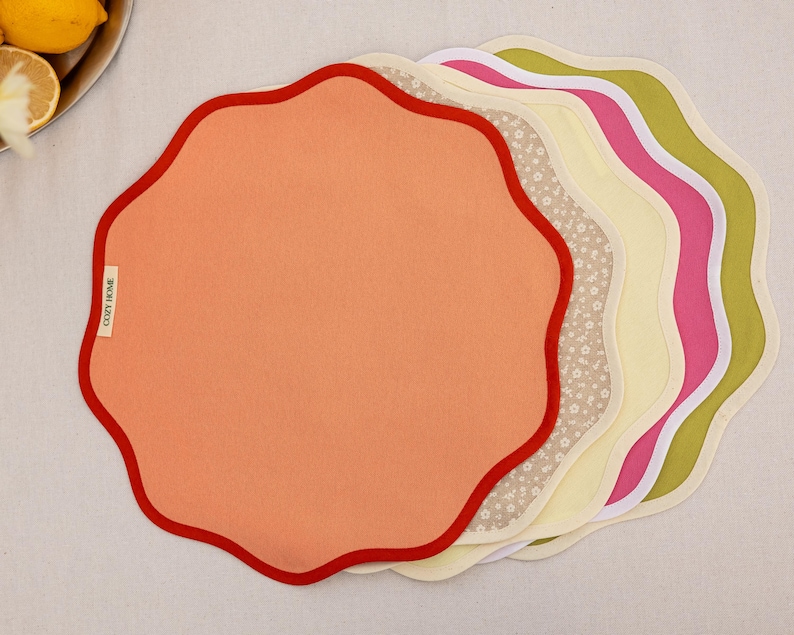 May include: A stack of colorful, scalloped placemats. The top placemat is peach with a red border. Other placemats include floral, yellow, pink, and green designs. The label on the peach placemat reads "COZY HOME".