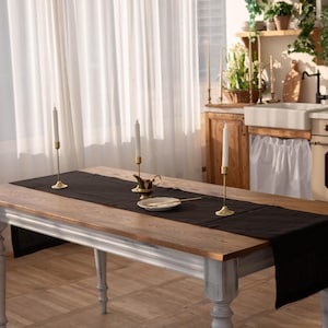 May include: A wooden dining table with a black table runner and gold candle holders. The table has a light wood top and gray painted legs. The room has white curtains and a kitchen in the background.