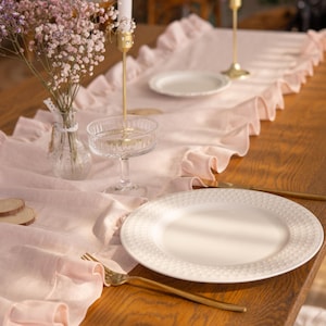 May include: A pink ruffled table runner with white plates, a glass, and candlesticks on a wooden table.
