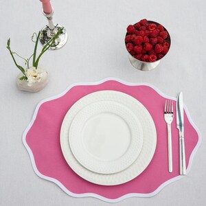 Op de afbeelding: Een gedekte tafel met een roze placemat, witte borden en bestek. Een kleine metalen kom is gevuld met verse frambozen. Een roze kandelaar en een klein bloemstuk staan ook op het witte tafelkleed.