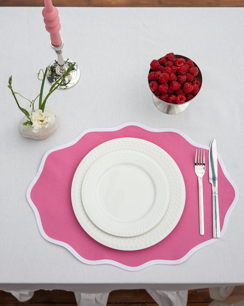 Op de afbeelding: Een gedekte tafel met een roze placemat, witte borden en bestek. Een kleine metalen kom is gevuld met verse frambozen. Een roze kandelaar en een klein bloemstuk staan ook op het witte tafelkleed.