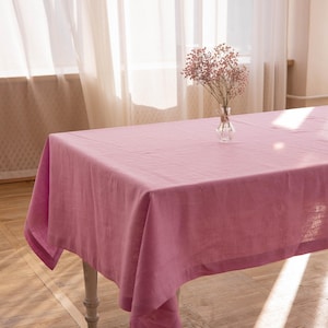 May include: A rectangular, dusty rose linen tablecloth draped over a white-legged table. A small glass vase with pink flowers sits on the table. The scene is illuminated by natural light from a nearby window.