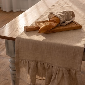 Könnte beinhalten: Ein beigefarbener Tischläufer aus Leinen mit einem gekräuselten Rand. Der Läufer liegt auf einem Holztisch mit einem Baguette auf einem Holzbrett.