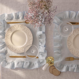 Puede incluir: Dos manteles individuales de color azul claro con bordes ondulados, cada uno con un plato de cer&aacute;mica blanca con un dise&ntilde;o decorativo, un cuchillo y un tenedor dorados y un vaso transparente.