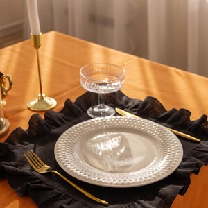 Puede incluir: Un mantel individual de lino negro con volantes con un plato blanco, un tenedor y un cuchillo dorados, y un vaso transparente sobre un mantel naranja.