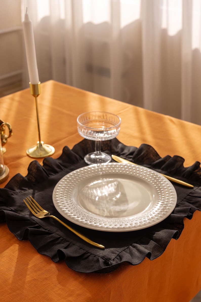 Puede incluir: Un mantel individual de lino negro con volantes con un plato blanco, un tenedor y un cuchillo dorados, y un vaso transparente sobre un mantel naranja.