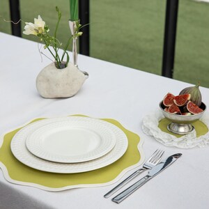 May include: A table setting with a white tablecloth, featuring a white plate set on a green placemat, silverware, and a small bowl of sliced figs. A small, white, hedgehog-shaped vase holds flowers.