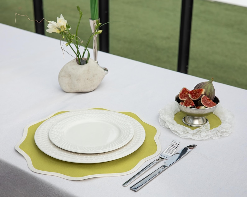 May include: A table setting with a white tablecloth, featuring a white plate set on a green placemat, silverware, and a small bowl of sliced figs. A small, white, hedgehog-shaped vase holds flowers.