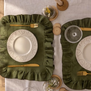 Puede incluir: Dos manteles individuales verdes con bordes festoneados, cada uno con un plato blanco y cubiertos de oro. Los manteles individuales est&aacute;n sobre un mantel de lino blanco.