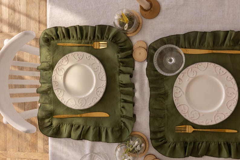 Puede incluir: Dos manteles individuales verdes con bordes festoneados, cada uno con un plato blanco y cubiertos de oro. Los manteles individuales est&aacute;n sobre un mantel de lino blanco.