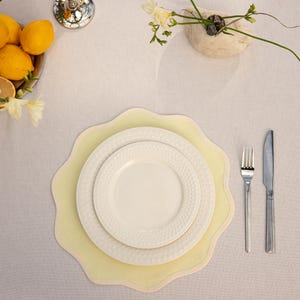 May include: Overhead shot of a table setting with a pale yellow placemat, white plates, and silverware. A small vase with flowers and a bowl of lemons are also on the table, creating a bright and inviting dining scene.