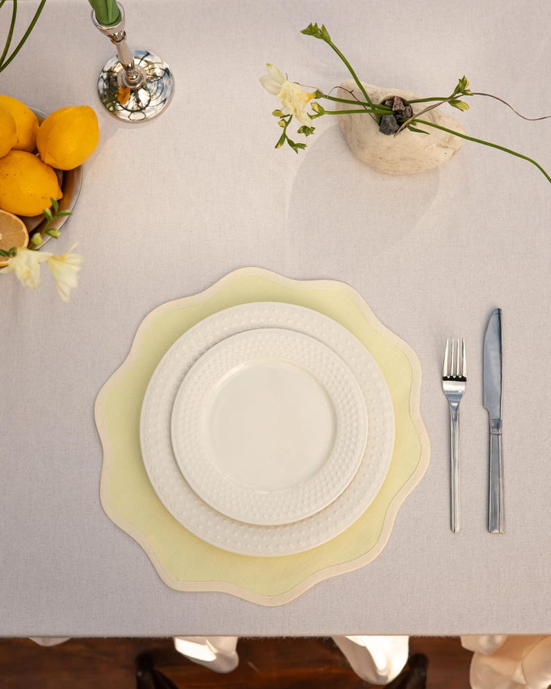 May include: Overhead shot of a table setting with a pale yellow placemat, white plates, and silverware. A small vase with flowers and a bowl of lemons are also on the table, creating a bright and inviting dining scene.