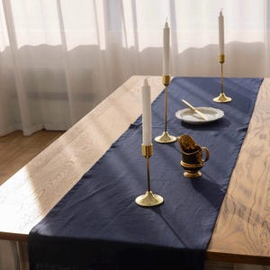 Puede incluir: Una mesa de madera está decorada con un camino de mesa azul marino, candelabros dorados con velas blancas, un plato pequeño con un cuchillo y una crema dorada y negra. La mesa está cerca de una cortina blanca transparente.