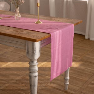 Peut inclure: Un chemin de table en lin rose sur une table en bois avec une base peinte en blanc. Un bougeoir doré avec une bougie blanche est sur le chemin de table.