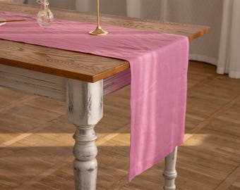 May include: A pink linen table runner on a wooden table with a white painted base. A gold candle holder with a white candle is on the table runner.