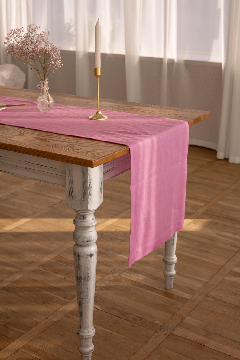 May include: A pink linen table runner on a wooden table with a white painted base. A gold candle holder with a white candle is on the table runner.