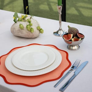 May include: A table setting with a white tablecloth, featuring white plates on an orange placemat with a red border. Silverware, a small bowl of figs, and a decorative stone with greenery complete the elegant arrangement.