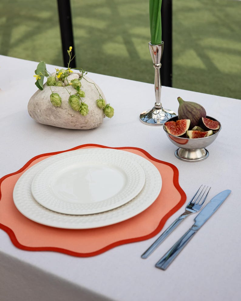 May include: A table setting with a white tablecloth, featuring white plates on an orange placemat with a red border. Silverware, a small bowl of figs, and a decorative stone with greenery complete the elegant arrangement.