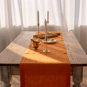 May include: A wooden table with a burnt orange linen table runner and three gold candle holders with white candles. A small gold creamer sits on the table runner.