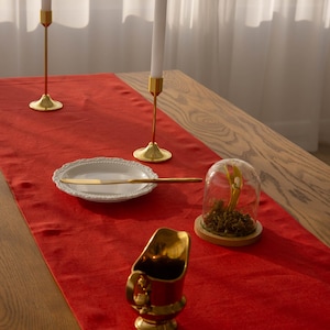 May include: A red table runner with gold candlesticks, a white plate with a gold knife, a glass cloche with dried flowers, and a gold pitcher.