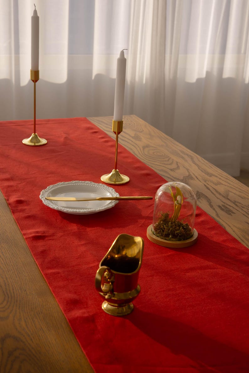 May include: A red table runner with gold candlesticks, a white plate with a gold knife, a glass cloche with dried flowers, and a gold pitcher.