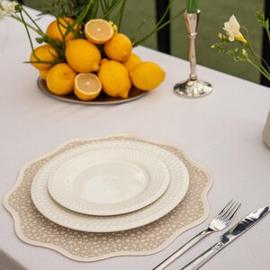 May include: A table setting featuring a beige floral placemat, two white plates, and silverware. A plate of lemons and flowers is in the background. A green candle sits in a silver candlestick.
