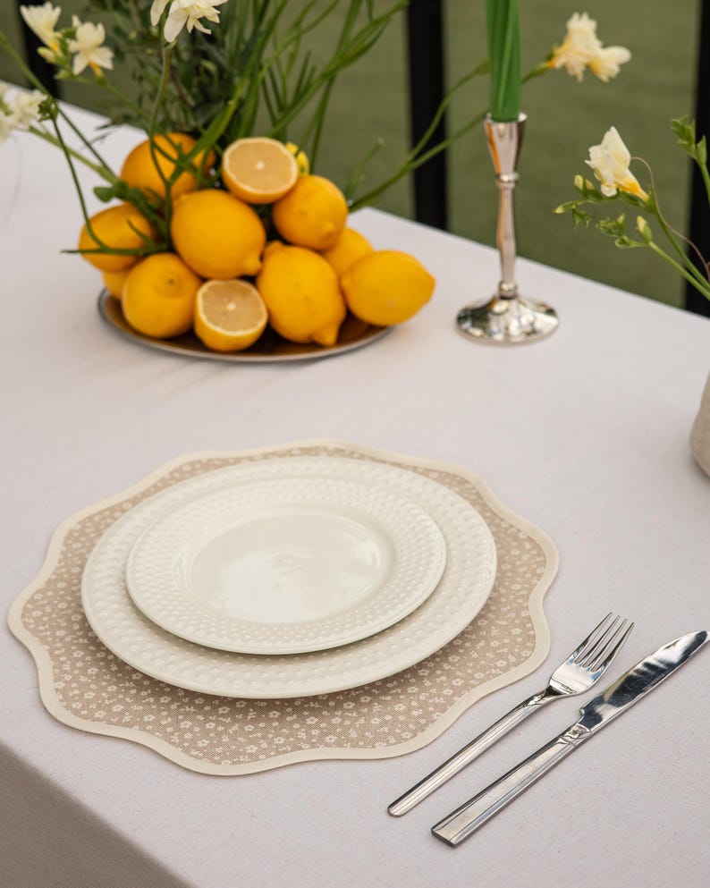 May include: A table setting featuring a beige floral placemat, two white plates, and silverware. A plate of lemons and flowers is in the background. A green candle sits in a silver candlestick.