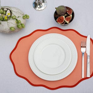 Op de afbeelding: Een tafelsetting met een koraalkleurige placemat met geschulpte rand, twee witte borden, bestek en een kleine kom met vijgen. Een decoratieve stenen kom met groen is ook aanwezig.