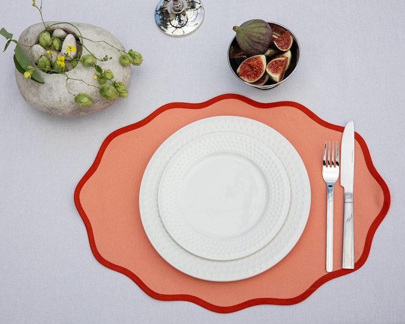 Op de afbeelding: Een tafelsetting met een koraalkleurige placemat met geschulpte rand, twee witte borden, bestek en een kleine kom met vijgen. Een decoratieve stenen kom met groen is ook aanwezig.
