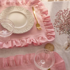 May include: Two pink ruffled linen placemats with white plates and gold silverware on a white linen tablecloth.