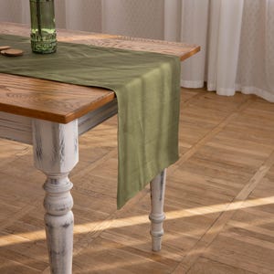 May include: A rustic wooden table with a distressed white finish and turned legs. A green linen table runner is draped across the table.