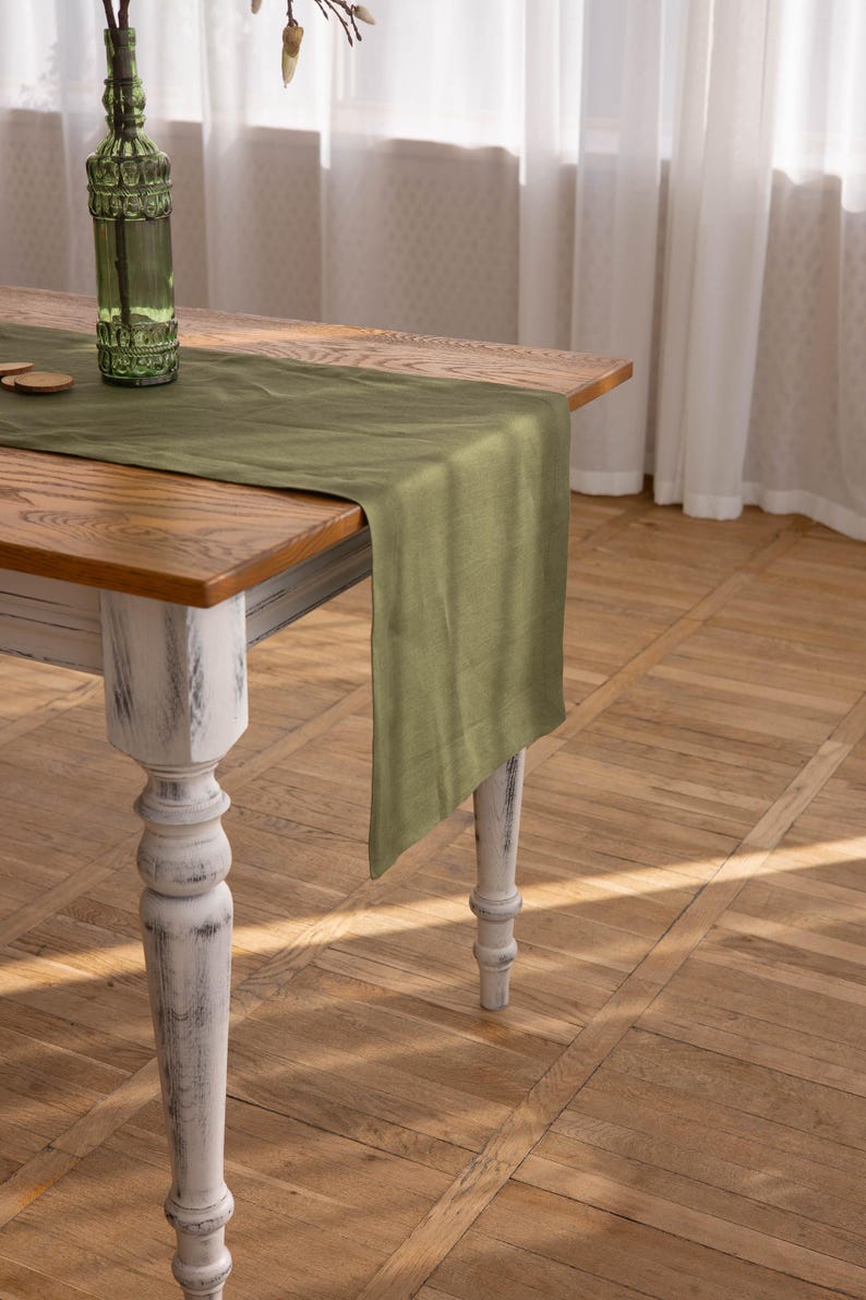 May include: A rustic wooden table with a distressed white finish and turned legs. A green linen table runner is draped across the table.