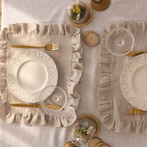 Puede incluir: Dos cubiertos con platos blancos con dise&ntilde;os en relieve, cubiertos de oro y vasos de vidrio transparentes. Los cubiertos est&aacute;n sobre manteles individuales de lino beige con bordes ondulados.