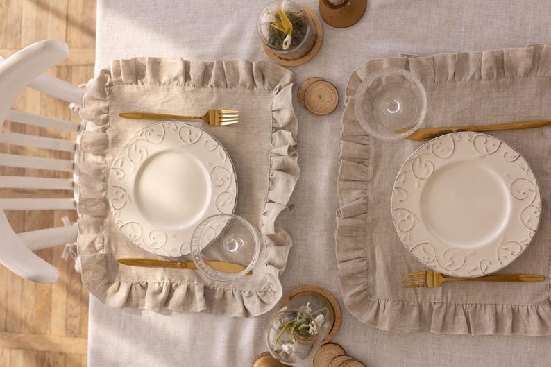 Puede incluir: Dos cubiertos con platos blancos con dise&ntilde;os en relieve, cubiertos de oro y vasos de vidrio transparentes. Los cubiertos est&aacute;n sobre manteles individuales de lino beige con bordes ondulados.