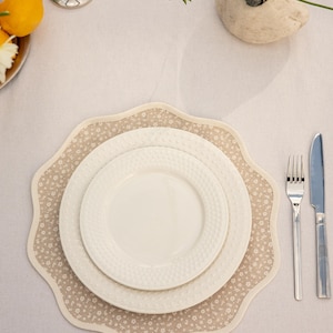 May include: A table setting featuring white plates on a floral placemat. The placemat has a scalloped edge and a beige background with small white flowers. Silverware, including a fork and knife, is placed to the right.