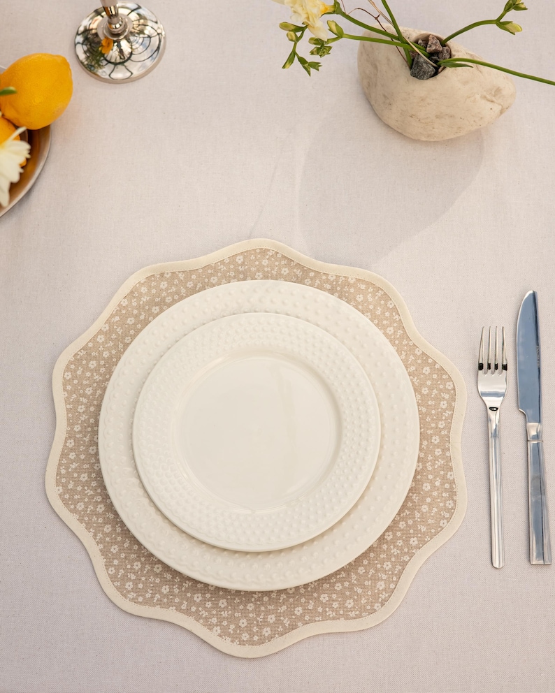 May include: A table setting featuring white plates on a floral placemat. The placemat has a scalloped edge and a beige background with small white flowers. Silverware, including a fork and knife, is placed to the right.