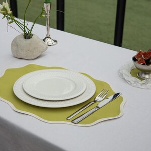 Op de afbeelding: Een tafelsetting met een wit tafelkleed, een lichtgroene placemat met witte rand en twee witte borden. Zilveren bestek ligt ernaast. Een kom met vijgen en een stenen vaas met bloemen zijn ook aanwezig.