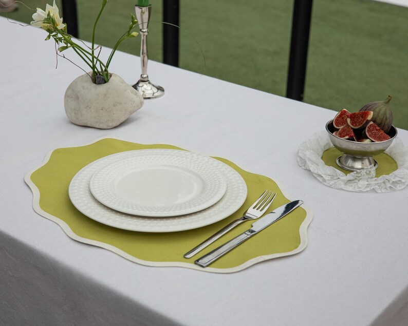 Op de afbeelding: Een tafelsetting met een wit tafelkleed, een lichtgroene placemat met witte rand en twee witte borden. Zilveren bestek ligt ernaast. Een kom met vijgen en een stenen vaas met bloemen zijn ook aanwezig.