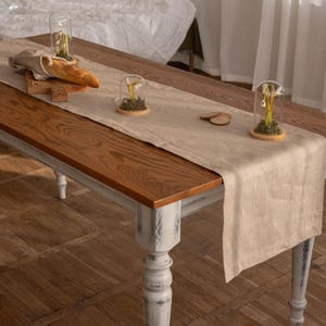 Könnte beinhalten: Ein rustikaler Holztisch mit weißer, abgenutzter Oberfläche und gedrehten Beinen. Ein beigefarbener Tischläufer aus Leinen liegt auf dem Tisch, darauf drei Glasglocken mit kleinen weißen Blumen.