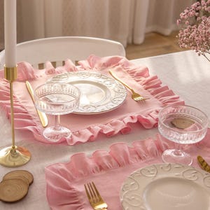 Puede incluir: Dos manteles individuales de color rosa con volantes, con platos blancos y cubiertos dorados. Los manteles individuales est&aacute;n sobre un mantel de lino blanco.